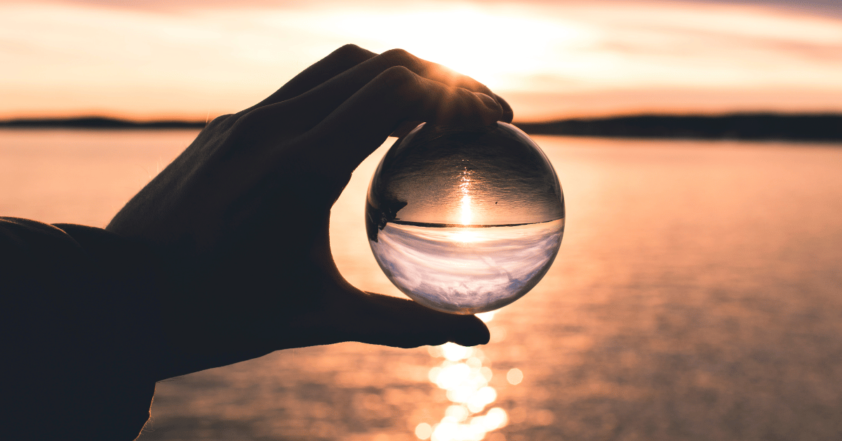 A person holding a sphere that is giving a reflection of a sunsetting on the ocean