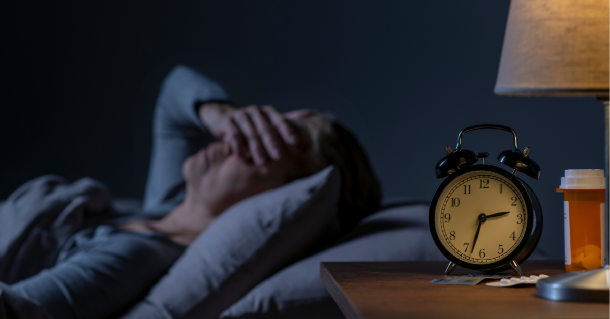 A man lying awake in bed with a bottle of prescription medication and an alarm clock on his nightstand