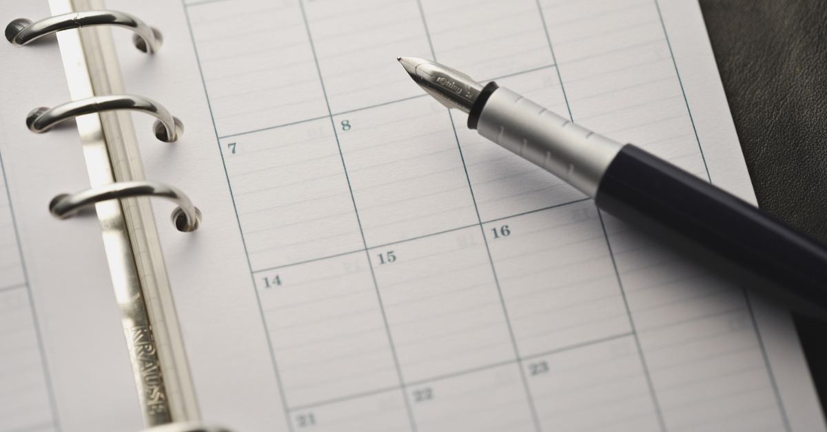 A fountain pen atop a paper notebook that is open to a month view of a calendar